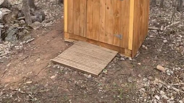 Una cabana de fusta amb un teulat inclinat i una porta de fusta, situada en una zona boscosa amb un camí de terra que hi condueix.