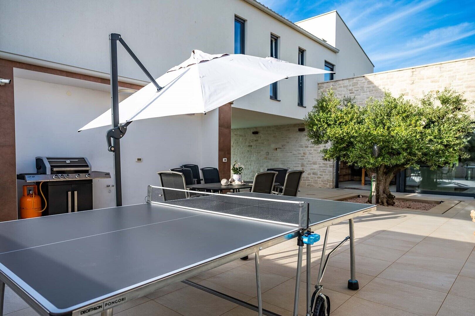 An outdoor table tennis table with a net, a grill, and chairs in a patio area.