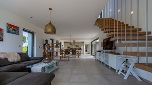 Un salon moderne avec un canapé, une table basse et des étagères. Un escalier mène à un espace cuisine comprenant une table à manger et des chaises.