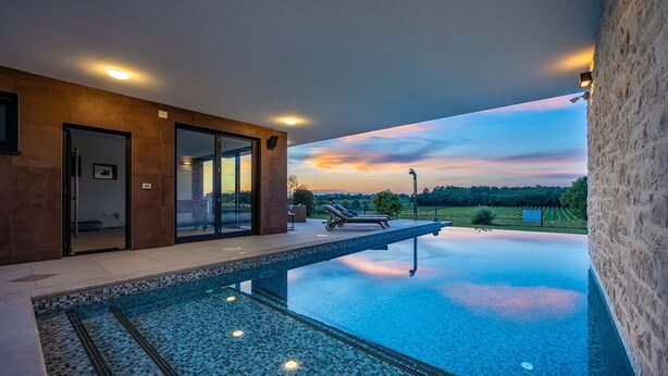 Un espace piscine extérieur moderne, doté d’une pièce fermée par des baies vitrées, d’un fauteuil de relaxation et offrant une vue sur un vignoble au coucher du soleil.
