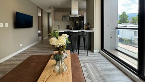 A modern hotel room with a large window, a flat-screen TV mounted on the wall, a kitchen area with a microwave, toaster, and coffee maker, and a wooden table with a vase of flowers.