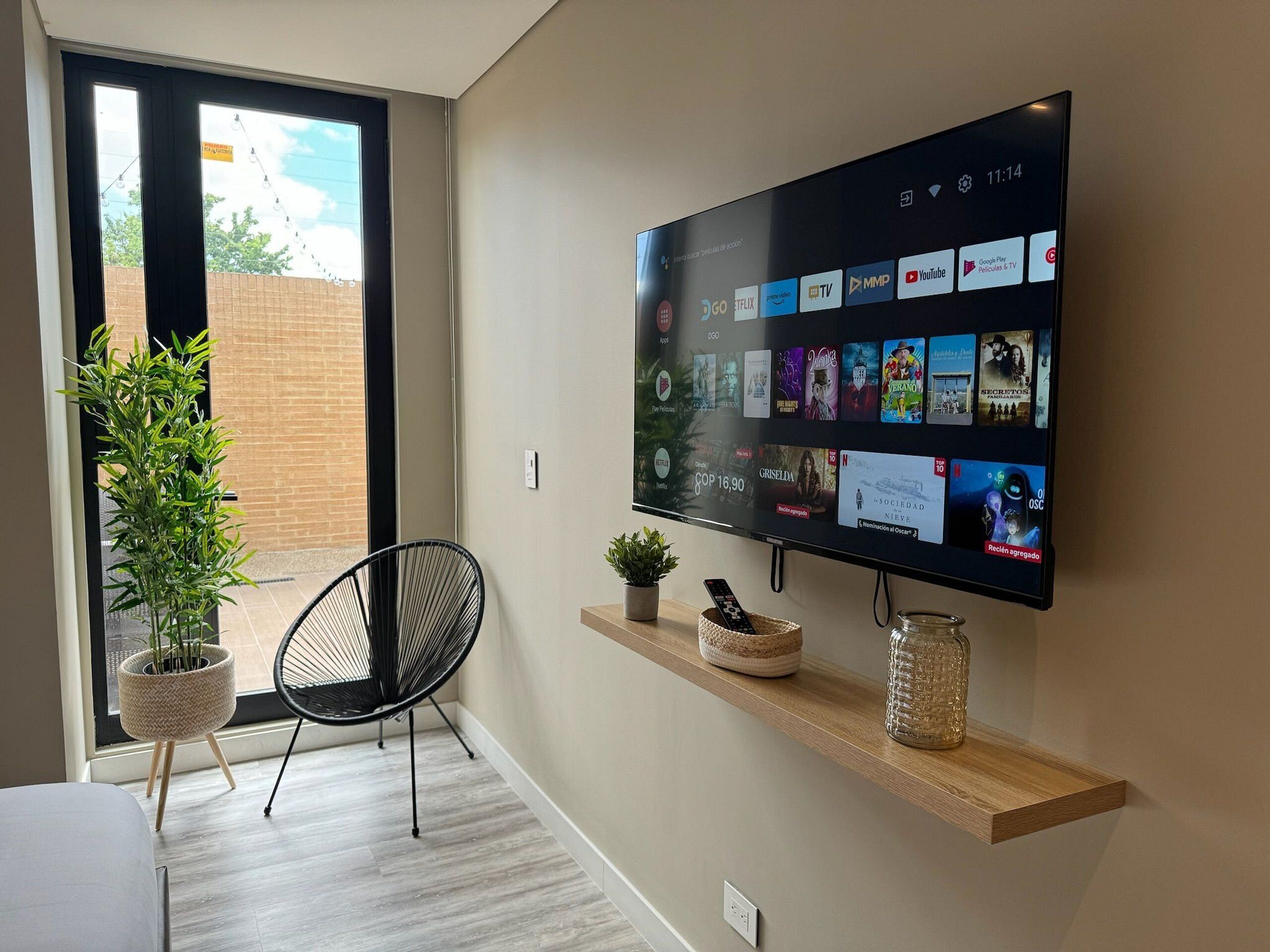 Un salon moderne avec un téléviseur à écran plat fixé au mur, qui diffuse différents services de streaming. On y trouve un fauteuil noir, une petite plante en pot et une étagère en bois ornée d’objets décoratifs.