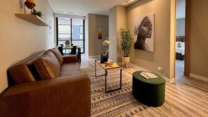 Een moderne woonkamer met een bruine leren bank, een salontafel en een groene poef. Er is een groot raam met uitzicht naar buiten, een schilderij aan de muur en een plant op een bijzettafeltje.