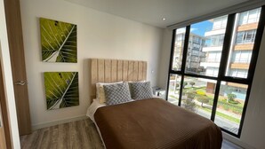 A modern bedroom with a large bed, two wall-mounted artworks, and a view of the city through large windows.