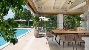 Un espace aménagé au bord de la piscine, avec une table et des chaises en bois, entouré de piliers en pierre et de parasols.