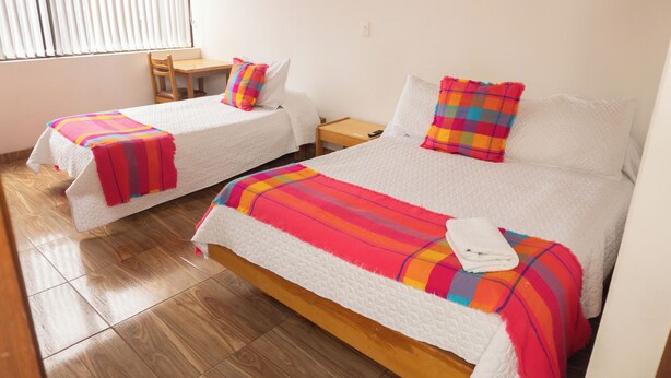 Habitación de hotel con dos camas, cada una con una manta y almohada coloridas, una mesita de noche de madera y una silla.