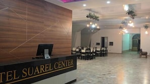Zona de recepción de hotel con mostrador de recepción identificado como "Hotel Suarel Center", suelo de baldosas y área de descanso con mesas y sillas.