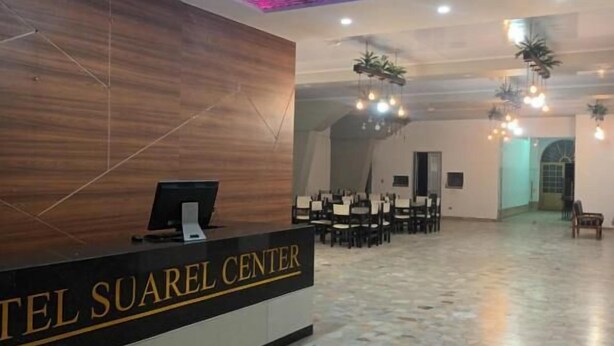 Zona de recepción de hotel con mostrador de recepción identificado como "Hotel Suarel Center", suelo de baldosas y área de descanso con mesas y sillas.