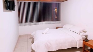 Un dormitorio con cama, mesita de noche, televisor montado en la pared y ventana con persianas verticales.
