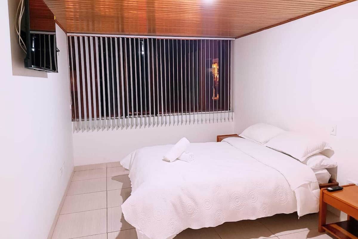 Ein Schlafzimmer mit einem Bett, einem Nachttisch, einem an der Wand montierten Fernseher und einem Fenster mit vertikalen Jalousien.
