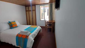 Un dormitorio con cama, una silla, una mesita, una ventana con cortinas y un televisor instalado en la pared.