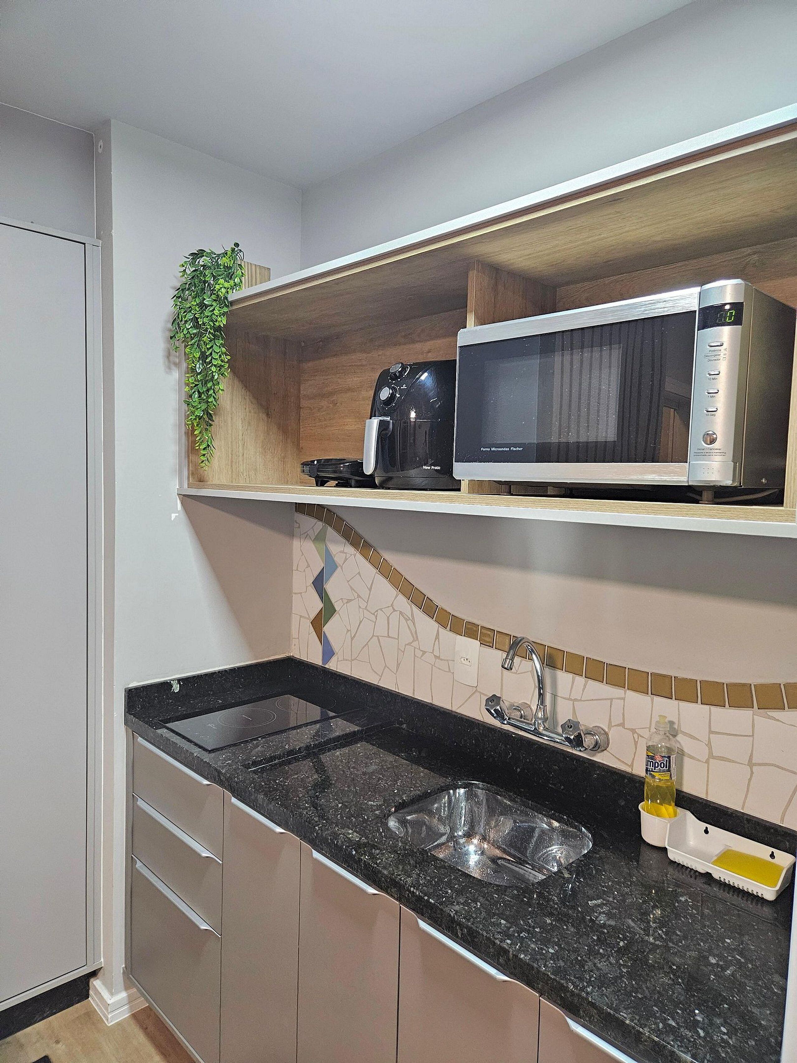 Uma cozinha com bancada preta, pia de aço inoxidável, micro-ondas e cafeteira.