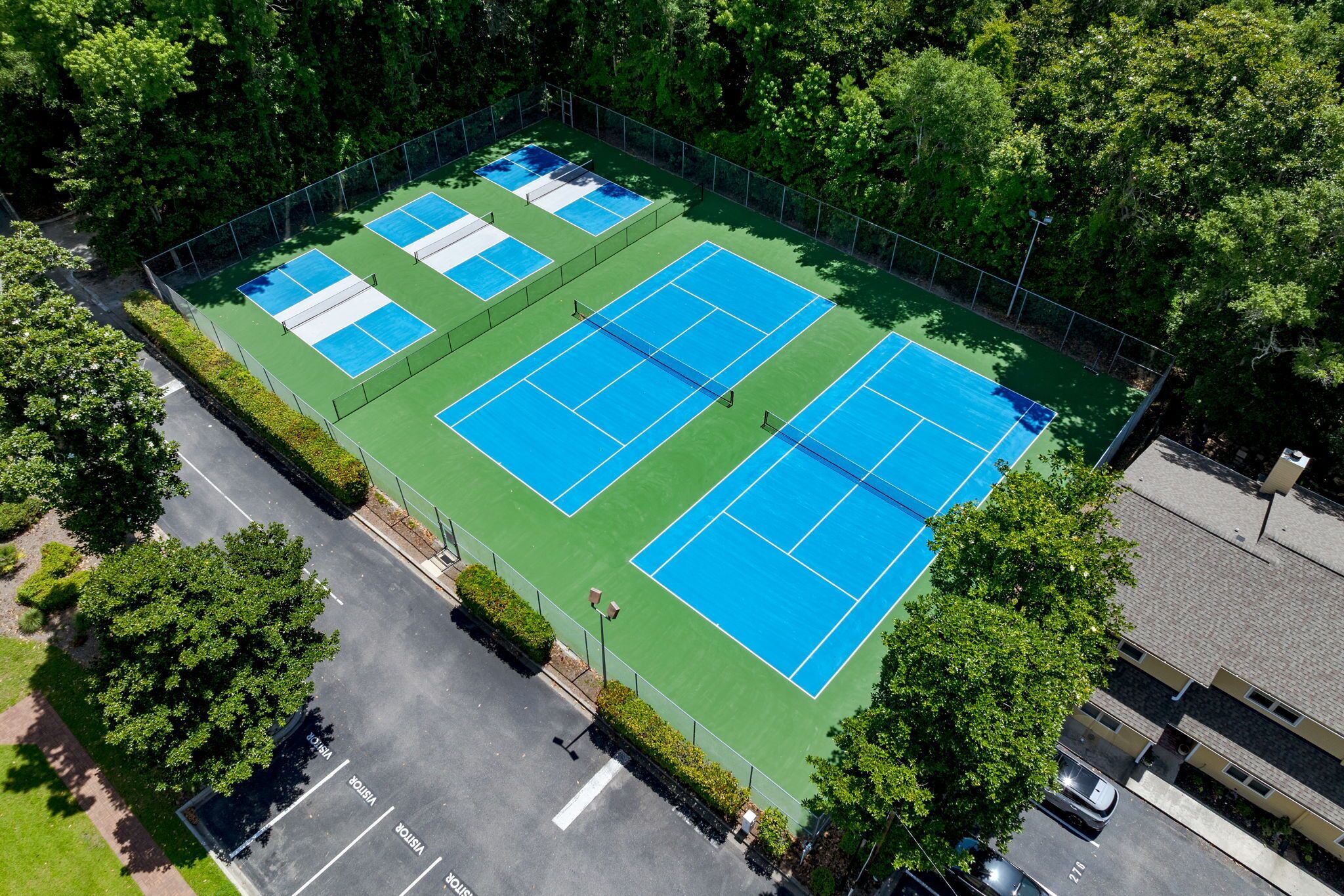 Vista aérea de un complejo de canchas de tenis con varias canchas, rodeado de árboles y junto a una carretera.