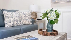 A living room with a grey sofa, patterned pillows, a vase with white flowers, and a book on a coffee table. - Updated/Resort StyleSleeps-8-Beach-5 min/Pool/Dining/pickle Ball-Prime location (Wilmington)