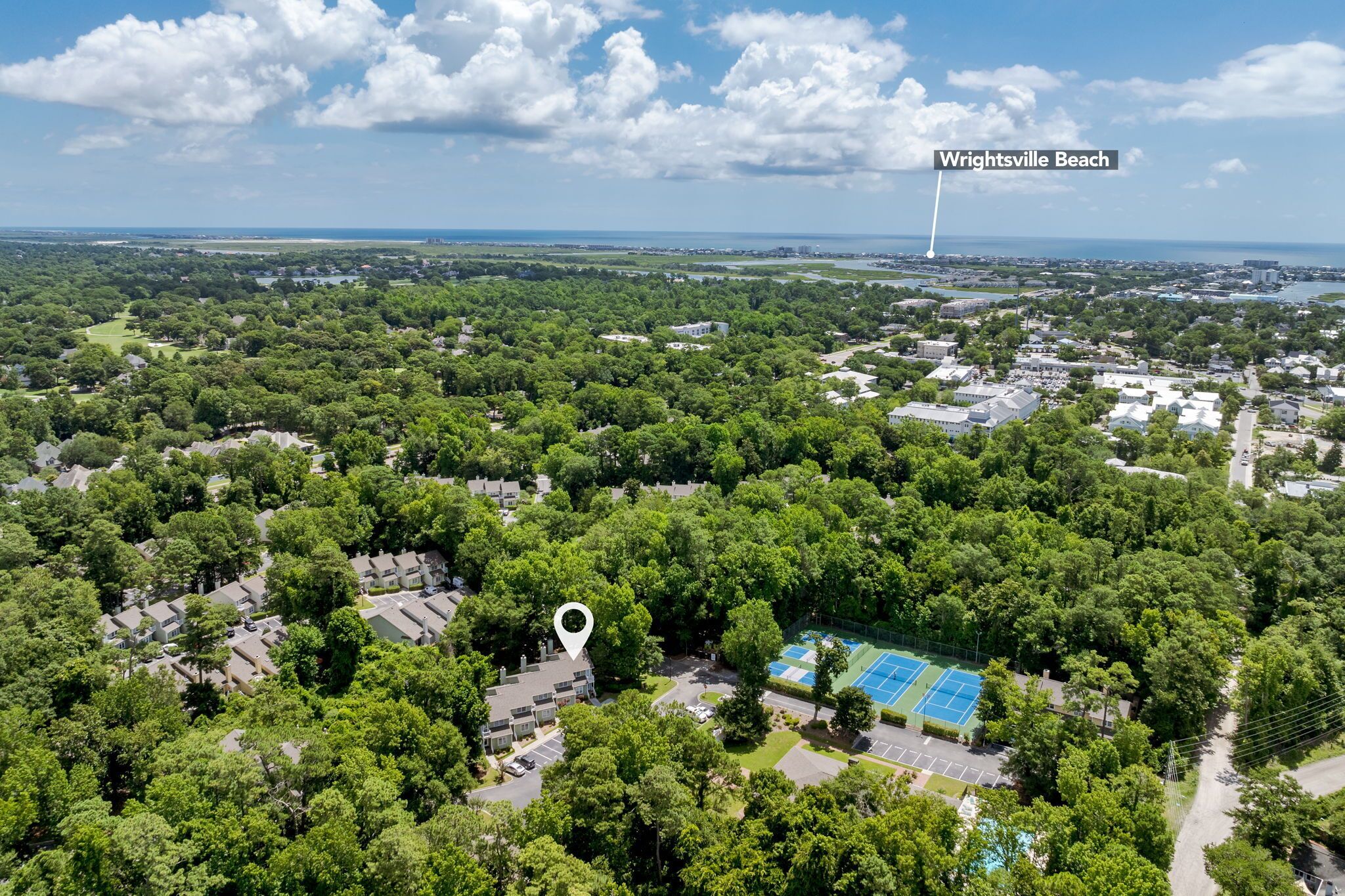 An aerial view of a residential area with a tennis court, surrounded by dense greenery and houses.
