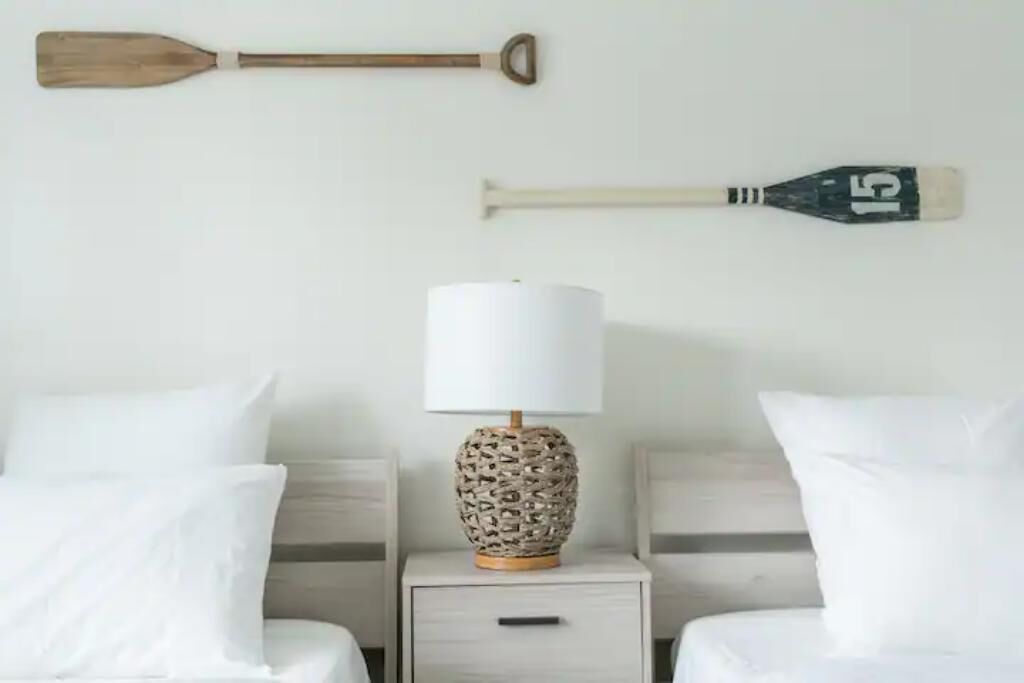 A neatly made bed with white bedding, a wooden paddle wall decoration, and a lamp with a patterned base on a bedside table.