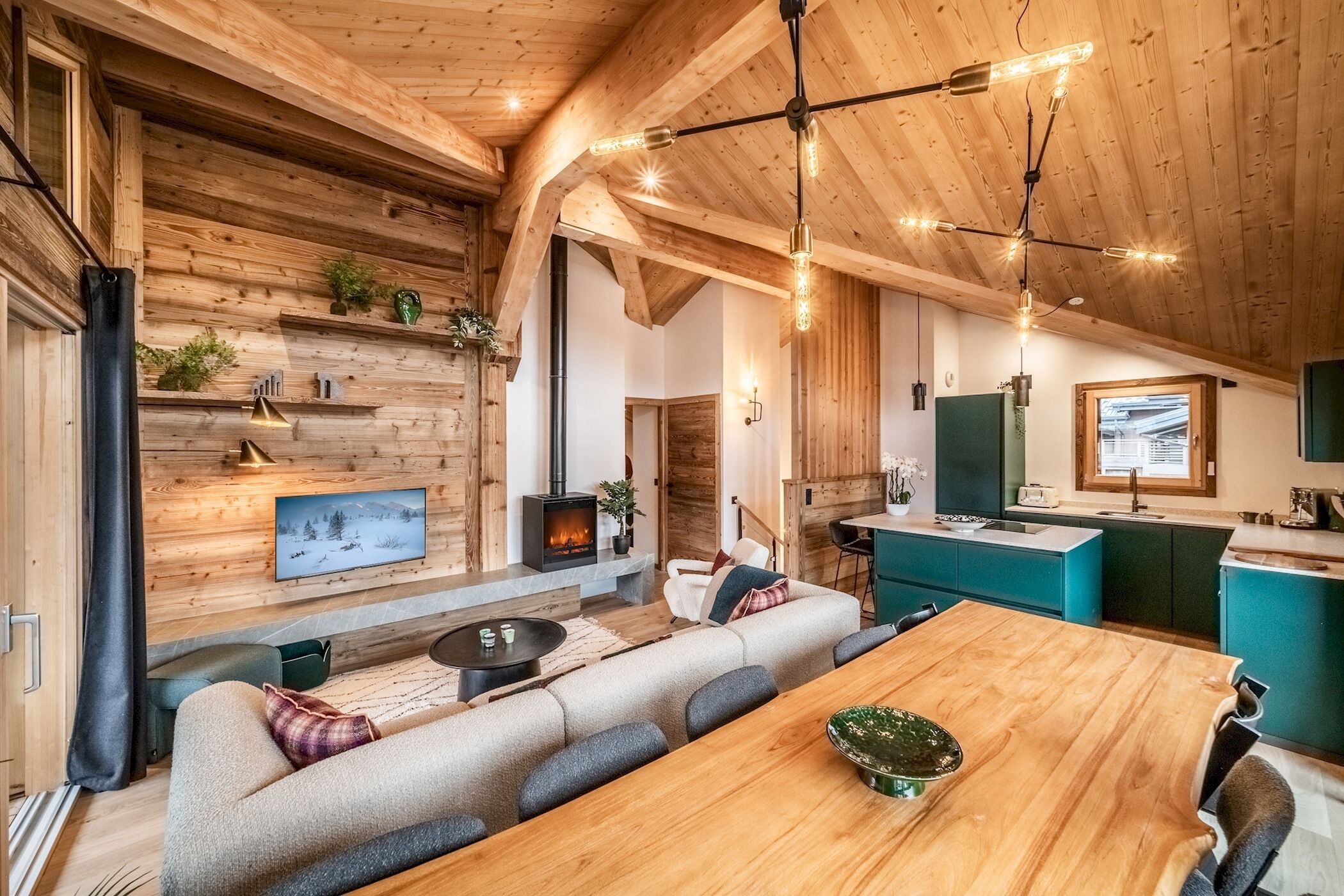 A cozy cabin interior with a wooden ceiling, a fireplace, a dining area, and a kitchen with teal cabinets.