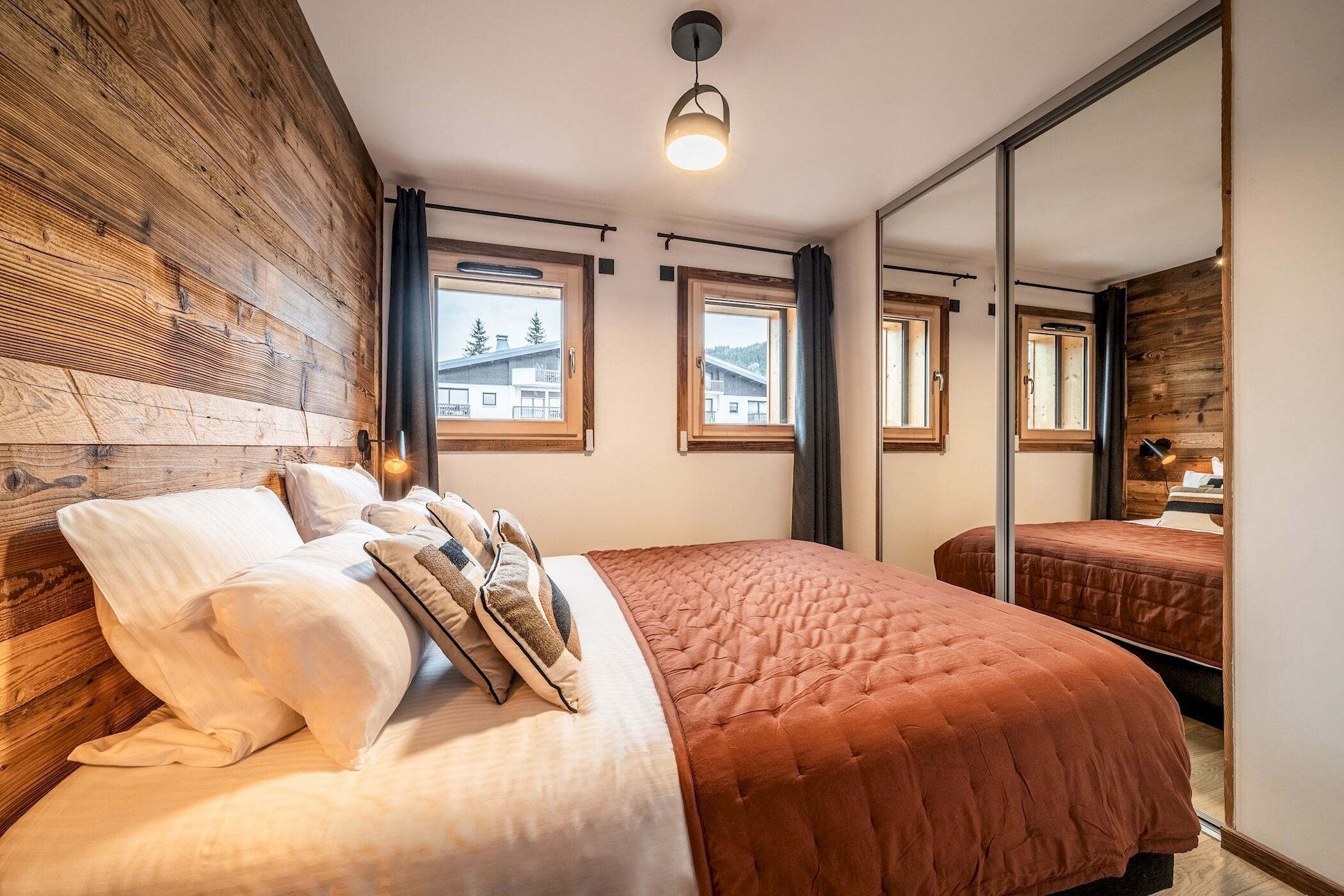 Une chambre avec un mur en bois, un lit avec une literie blanche et marron, et trois fenêtres avec des rideaux.