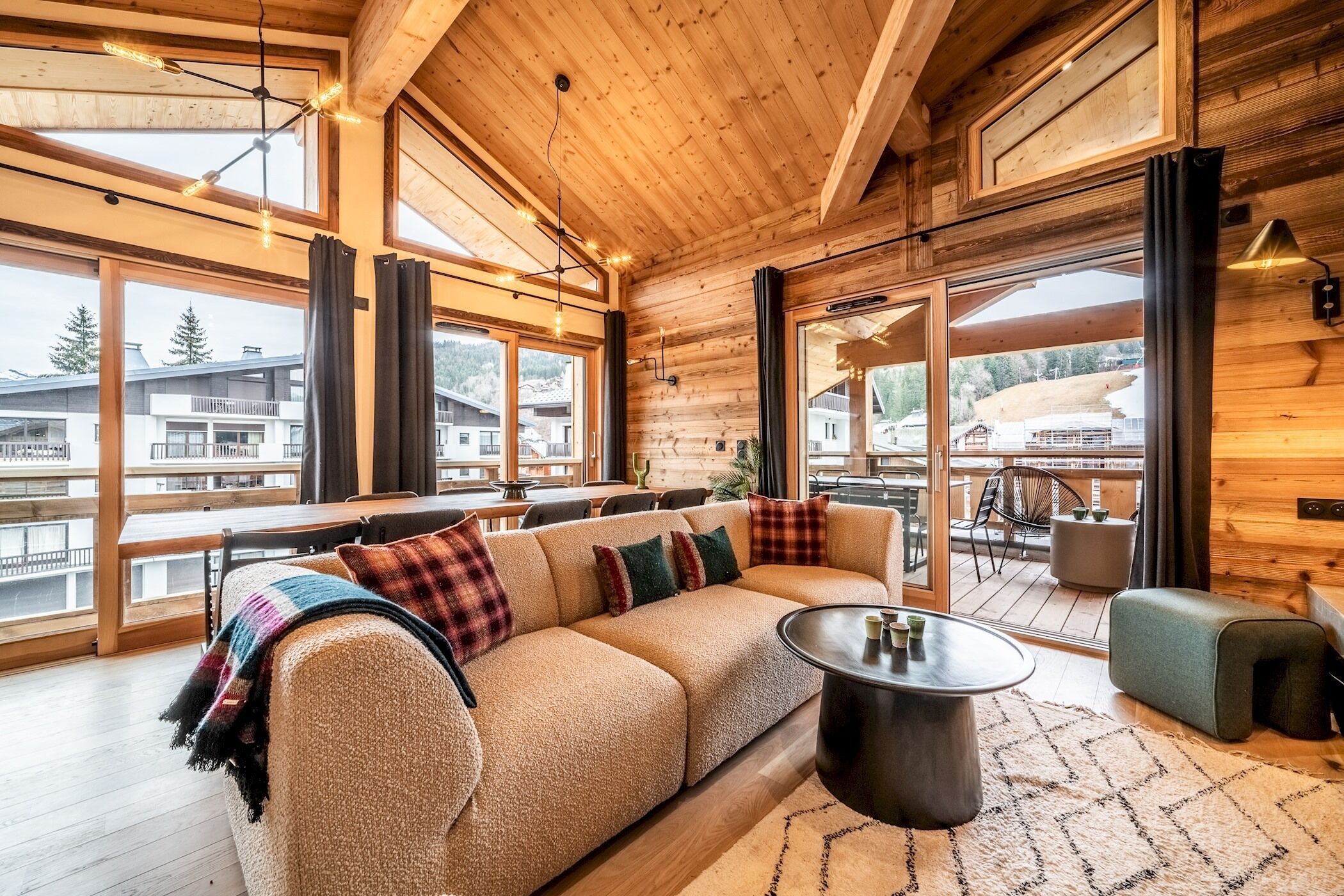 A cozy log cabin interior with a large sofa, a round coffee table, and a view of the surrounding landscape through large windows.