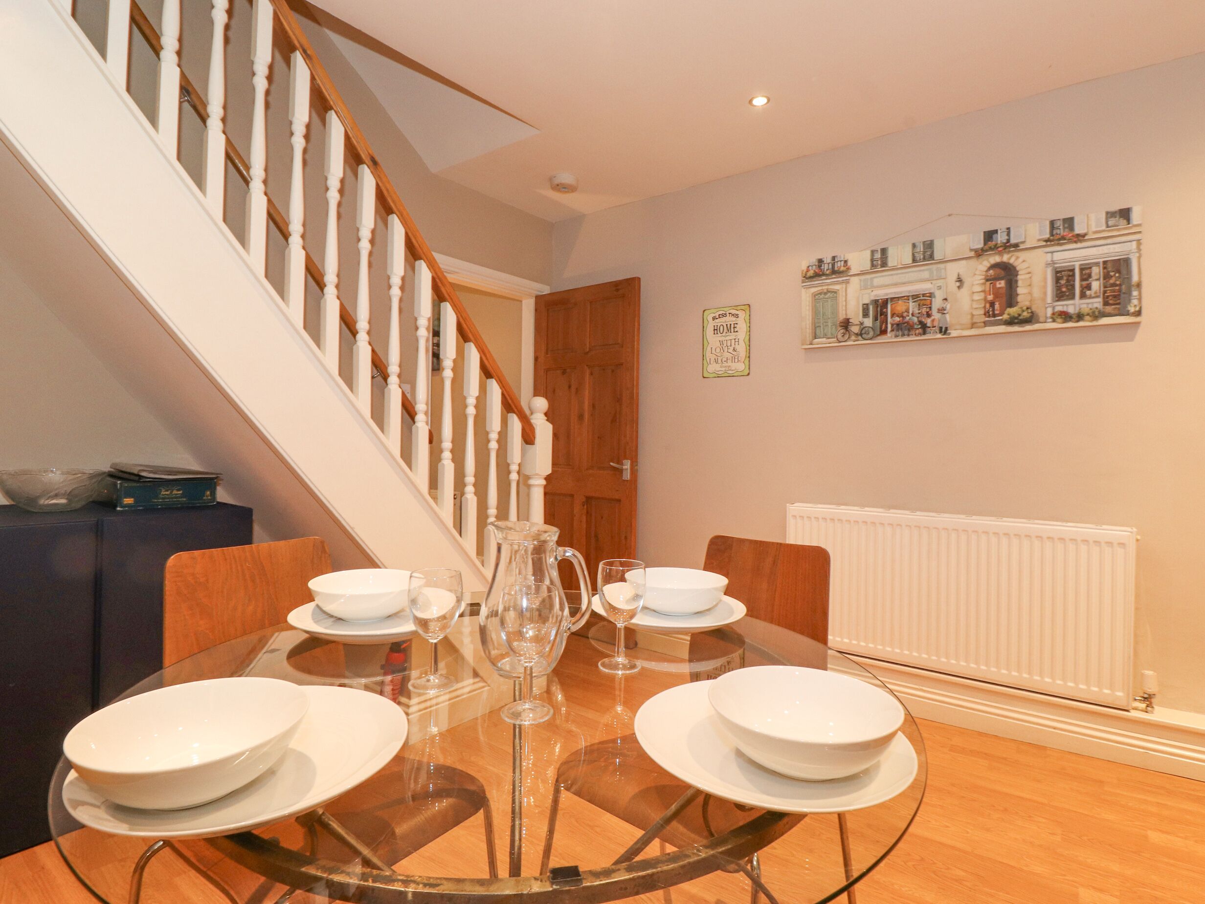 A dining area with a round glass table set for four, a wooden staircase, and a wall-mounted shelf with figurines.