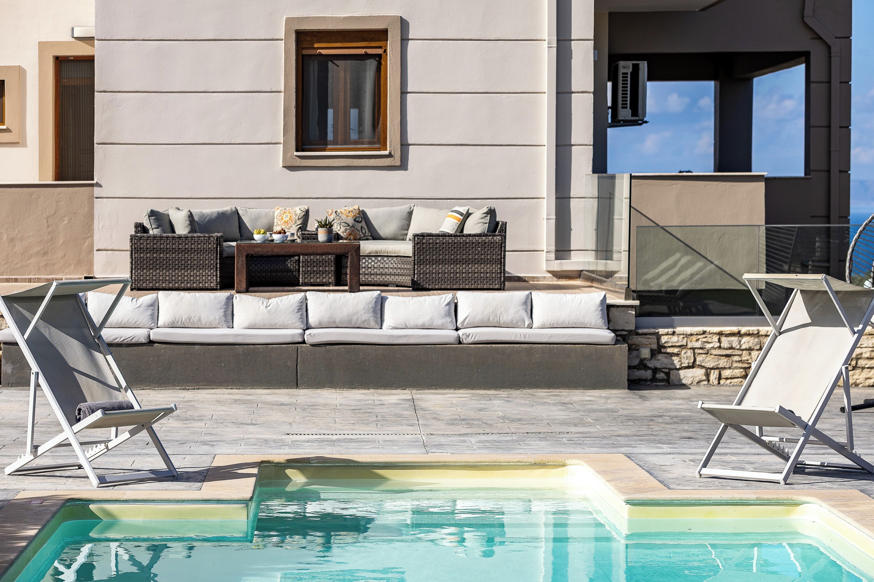 Un área junto a la piscina con un sofá y dos sillones reclinables, una mesita con adornos, y vista despejada a la piscina.