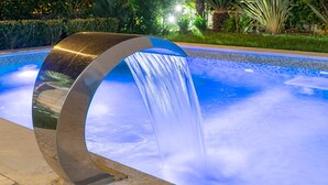 Una fuente moderna con una estructura metálica curva y un flujo de agua en cascada en un área de piscina rodeada de vegetación.