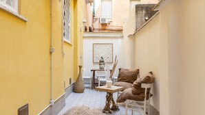 Une ruelle étroite aux murs jaunes, un sol carrelé de blanc, une petite table ornée d’un vase et de bougies, et une chaise avec des coussins.