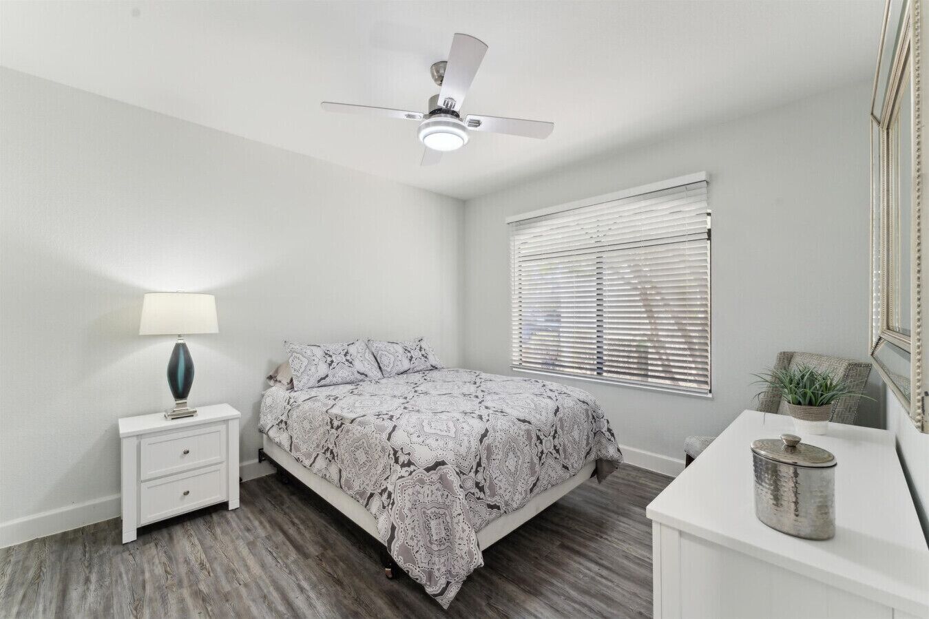 Un dormitorio con cama, mesita de noche con lámpara, cómoda, ventilador de techo y ventana con persianas.