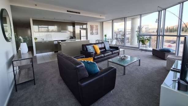 Ein modernes Wohnzimmer mit einer Couch, einem Couchtisch und einem Fernseher. Im Hintergrund befindet sich eine Küche mit einem großen Fenster.