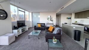 Ein modernes Wohnzimmer mit Sofa, Couchtisch und Fernseher. Es gibt eine Küchenzeile mit Spüle und Schränken.
