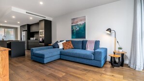 A modern living room with a blue sectional sofa, a kitchen area, and a large abstract painting on the wall. - Contemporary Tri-Level APT in a Prime Central Spot (Braddon)
