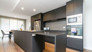 A modern kitchen with dark cabinetry, a central island, stainless steel appliances, and a dining area with a table and chairs. - Contemporary Tri-Level APT in a Prime Central Spot (Braddon)
