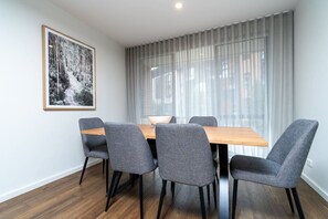 Un coin repas avec une table en bois et des chaises à revêtement gris, une photographie encadrée accrochée au mur et une grande fenêtre ornée de rideaux légers.