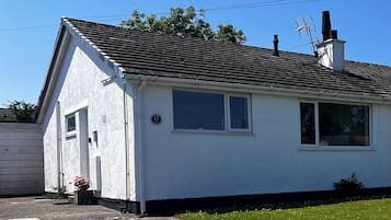 Una casa pequeña de un solo piso, de color blanco, con techo oscuro, una chimenea y un pequeño jardín con césped y una maceta.