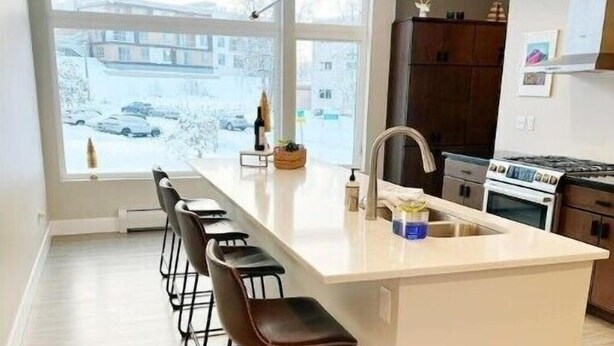 A modern kitchen with a large island, bar stools, a built-in oven, and a view of the outside. - Bootlegger’s Nest
 (Anchorage)