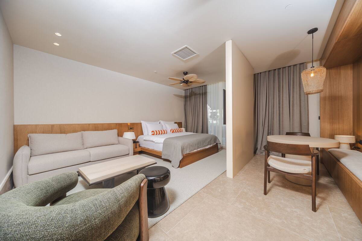 A modern hotel room with a sofa, a bed with orange pillows, a dining table, a chair, a small stool, and a ceiling fan.