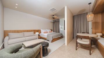 A modern hotel room with a sofa, a bed with orange pillows, a dining table, a chair, a small stool, and a ceiling fan.