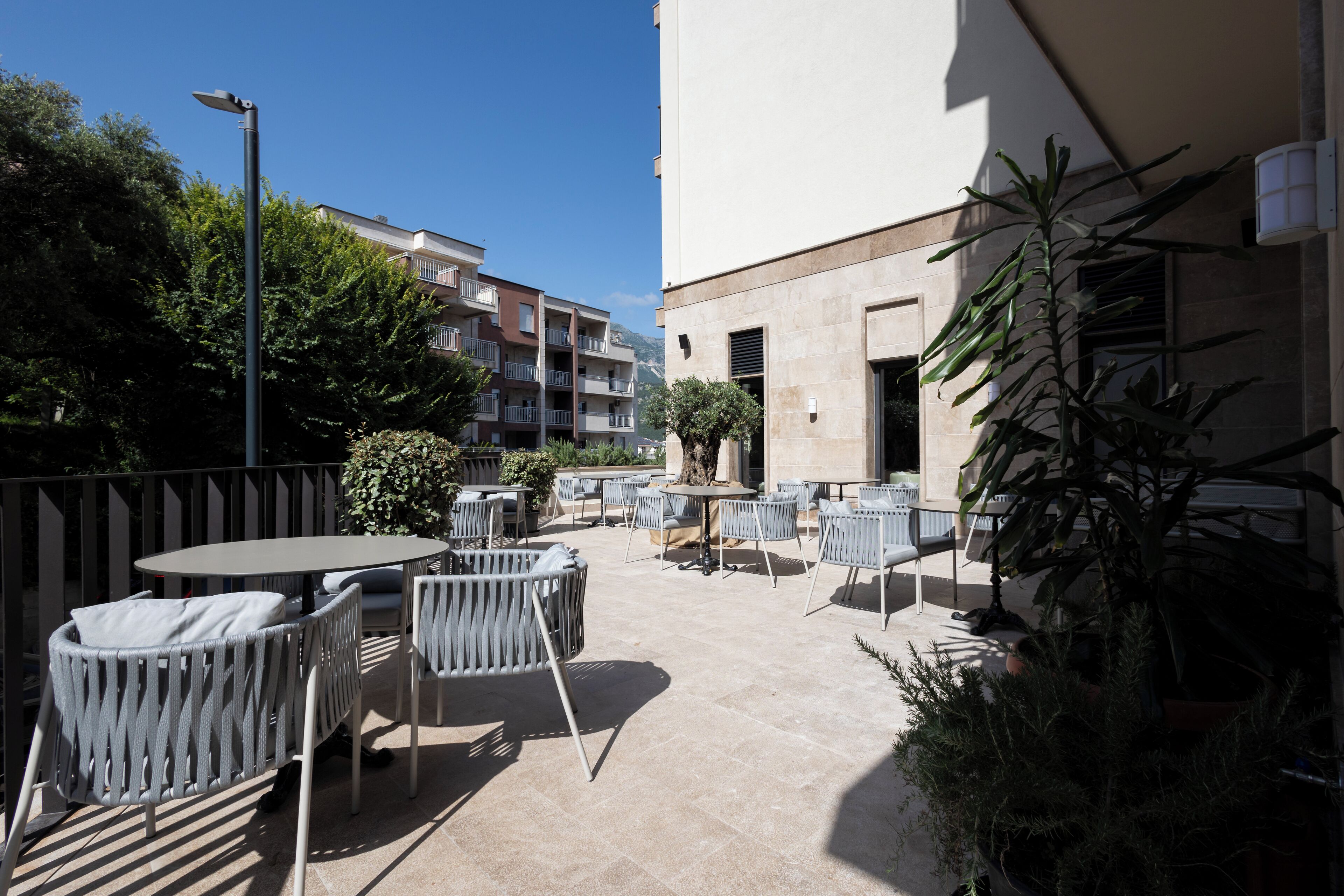 An outdoor patio with round tables and chairs, surrounded by potted plants and a building in the background.