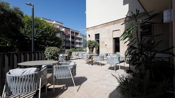 Eine Terrasse im Freien mit runden Tischen und Stühlen, umgeben von Kübelpflanzen und einem Gebäude im Hintergrund.