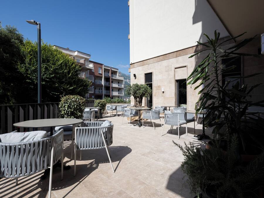 Eine Terrasse im Freien mit runden Tischen und Stühlen, umgeben von Kübelpflanzen und einem Gebäude im Hintergrund.