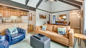 Ein geräumiger Wohnbereich mit einem Ledersofa, einem blauen Sessel und einem Couchtisch. Im Hintergrund befindet sich eine Küche mit Holzausbau und einem Kühlschrank.