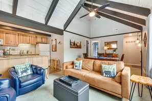 Ein geräumiger Wohnbereich mit einem Ledersofa, einem blauen Sessel und einem Couchtisch. Im Hintergrund befindet sich eine Küche mit Holzausbau und einem Kühlschrank.