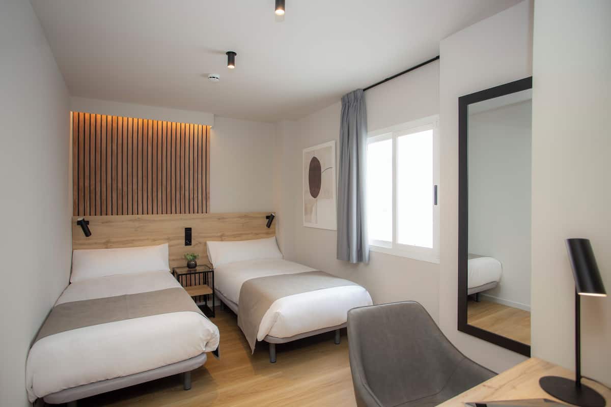 Una habitación de hotel moderna con dos camas, un escritorio, una silla, un espejo y una ventana con cortinas.