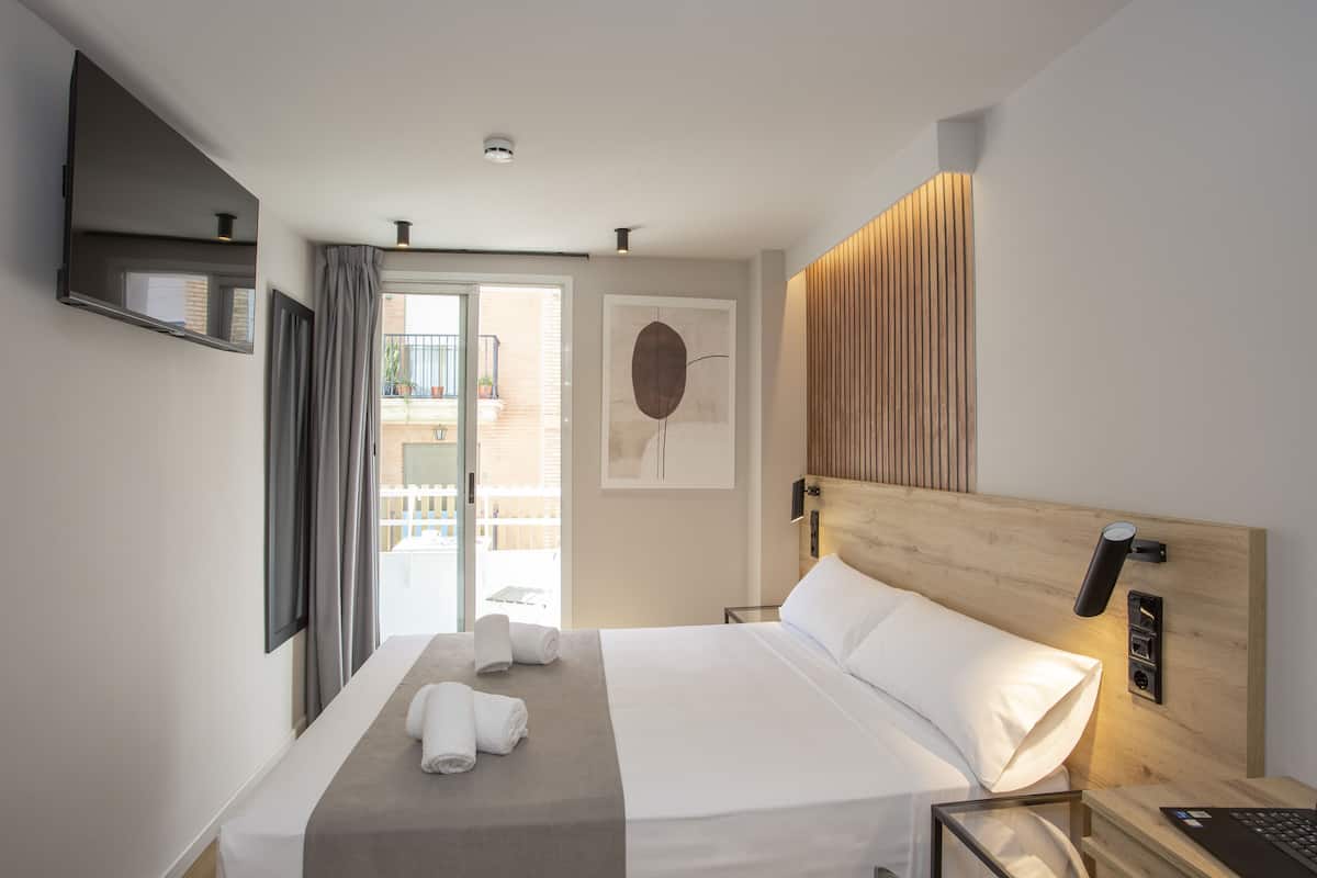 Una habitación de hotel moderna con una cama grande, un televisor de pantalla plana montado en la pared, un cabecero de madera y vistas al balcón exterior.