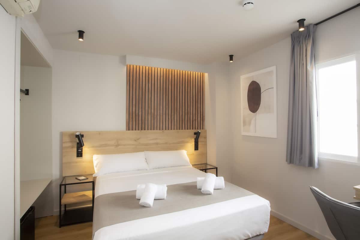 Una habitación de hotel moderna con un cabecero de madera, una cama grande, dos mesitas de noche, un escritorio y una ventana con cortinas.