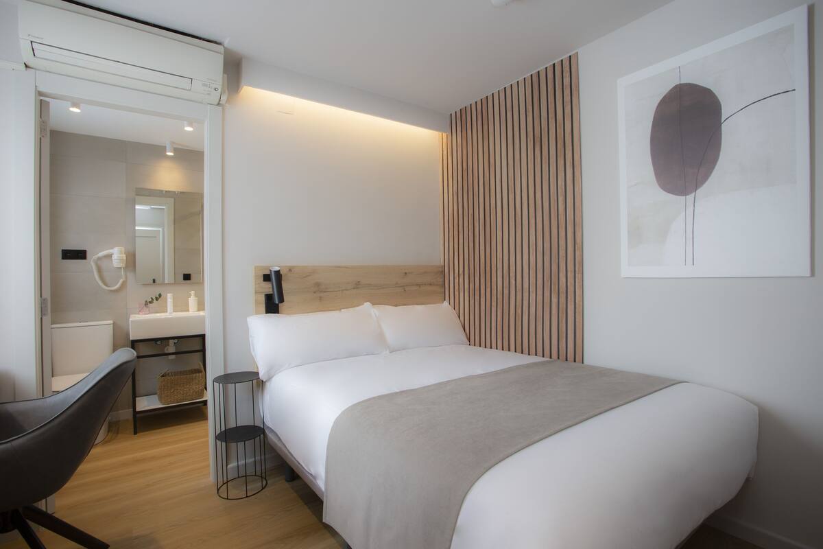 Una habitación de hotel moderna con cama, mesita de noche, silla y un ventanal grande.