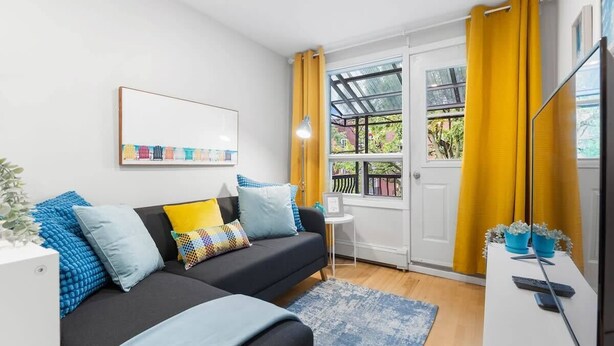 A modern living room with a black sofa, blue and yellow cushions, a small side table, and a large window with yellow curtains.