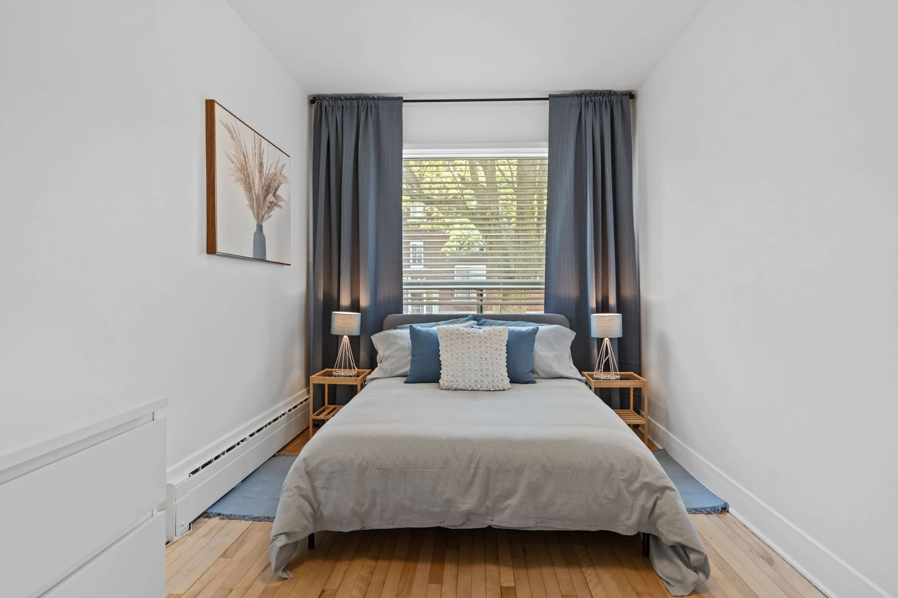 A bedroom with a large bed, two bedside tables with lamps, a window with curtains, and a framed picture on the wall.