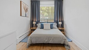 A bedroom with a large bed, two bedside tables with lamps, a window with curtains, and a framed picture on the wall. - All-Inclusive Furnished Large 2BR Apartment in Rosemont (Montréal)