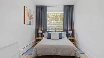 Une chambre à coucher avec un grand lit, deux tables de chevet avec des lampes, une fenêtre avec des rideaux et un tableau encadré au mur.
